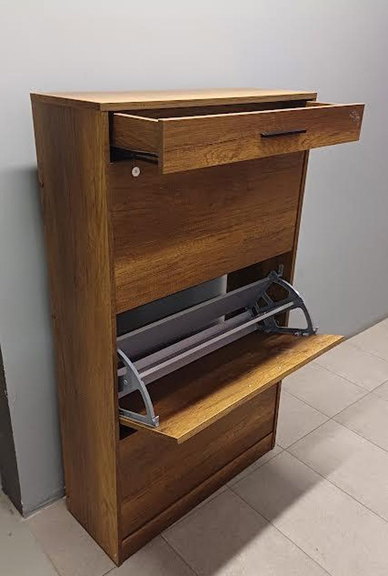 Op de afbeelding: Een bruine houten schoenenkast met een uittrekbare lade en twee planken. De kast heeft een uittrekbaar schoenenrek van grijs metaal.