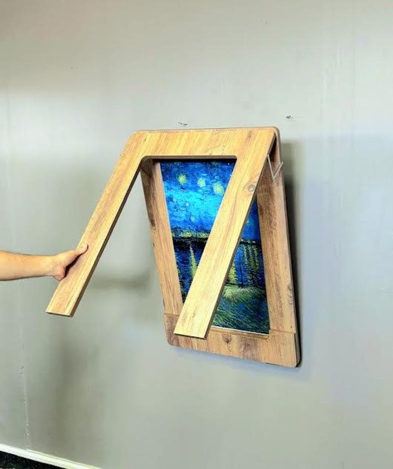 K&ouml;nnte beinhalten: Ein klappbarer Holztisch mit einem Gem&auml;lde der Sternennacht im Rahmen. Der Tisch ist zusammengeklappt und an einer Wand montiert.
