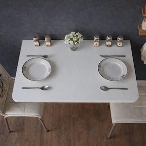 May include: Overhead shot of a small white table set for two. Each place setting includes a plate, spoon, and knife. A small vase of white flowers sits in the center, with spice jars and chairs on either side. The chairs have floral designs.