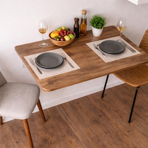 May include: A small, wooden dining table with a dark finish, set for two. The table has a bowl of fruit, wine glasses, and a small potted plant. Two chairs, one upholstered in gray fabric and the other woven, complete the dining set.