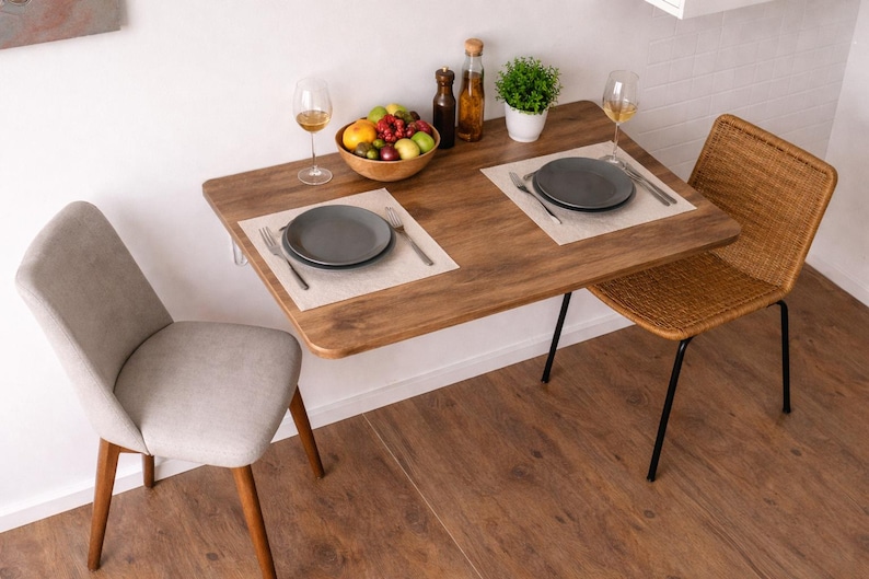 May include: A small, wooden dining table with a dark finish, set for two. The table has a bowl of fruit, wine glasses, and a small potted plant. Two chairs, one upholstered in gray fabric and the other woven, complete the dining set.