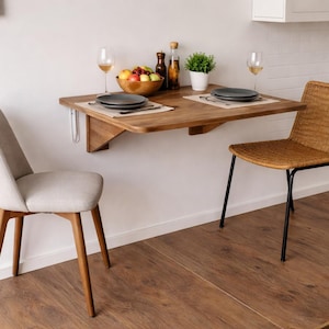May include: A wooden, wall-mounted dining table with a rounded edge, set with plates, placemats, and wine glasses. Two chairs, one upholstered in light gray fabric and the other woven, are positioned on either side. A bowl of fruit and condiments are on the table.