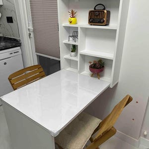 May include: White folding table with a white shelf unit attached to the wall. The shelf unit has three shelves and a small radio on the top shelf. There are two wooden chairs with beige cushions.