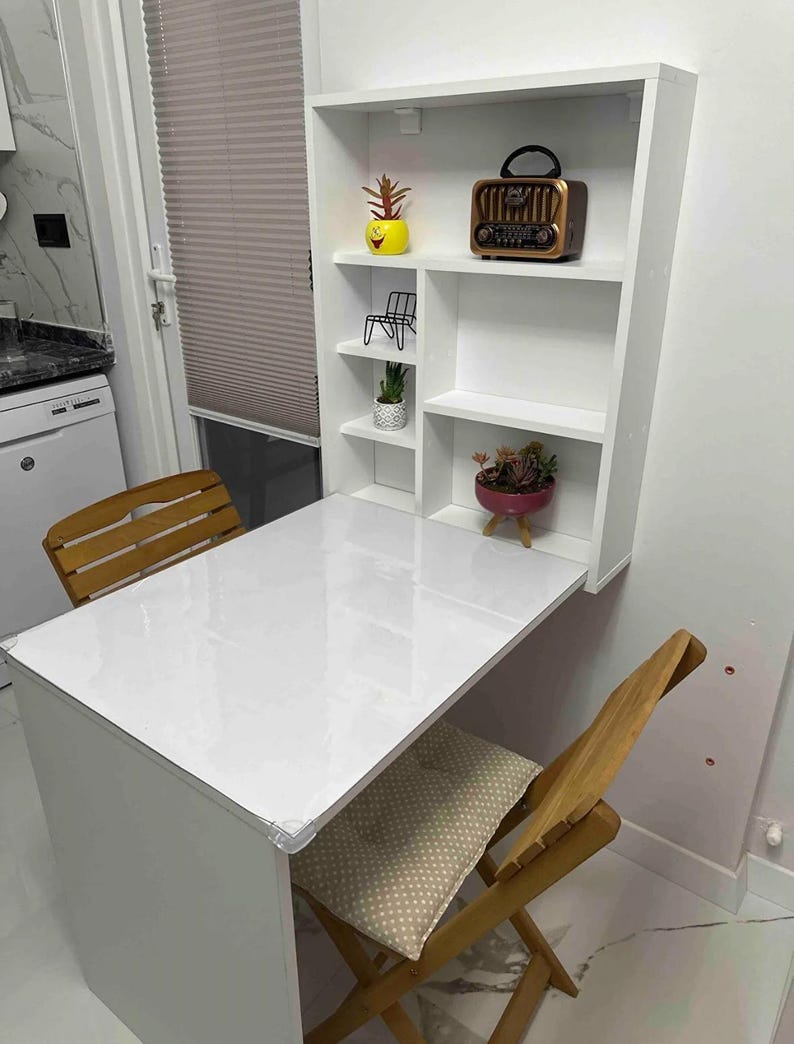 May include: White folding table with a white shelf unit attached to the wall. The shelf unit has three shelves and a small radio on the top shelf. There are two wooden chairs with beige cushions.