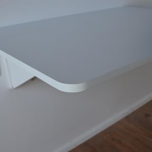 May include: A white floating shelf mounted on a white wall. The shelf has a rounded edge and is supported by a white bracket. The shelf is approximately 24 inches wide and 12 inches deep.