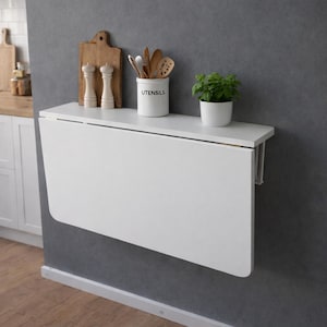 May include: A white, wall-mounted, folding table in a kitchen setting. The table is rectangular and has a clean, modern design. On the table are a small potted plant, a utensil holder, and decorative items. The kitchen has a neutral color scheme.