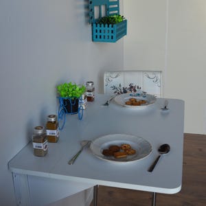 May include: A small, white table with two plates of cookies and silverware. A blue decorative planter with greenery sits on the table, along with spice jars. A blue wall-mounted planter with greenery is above the table.