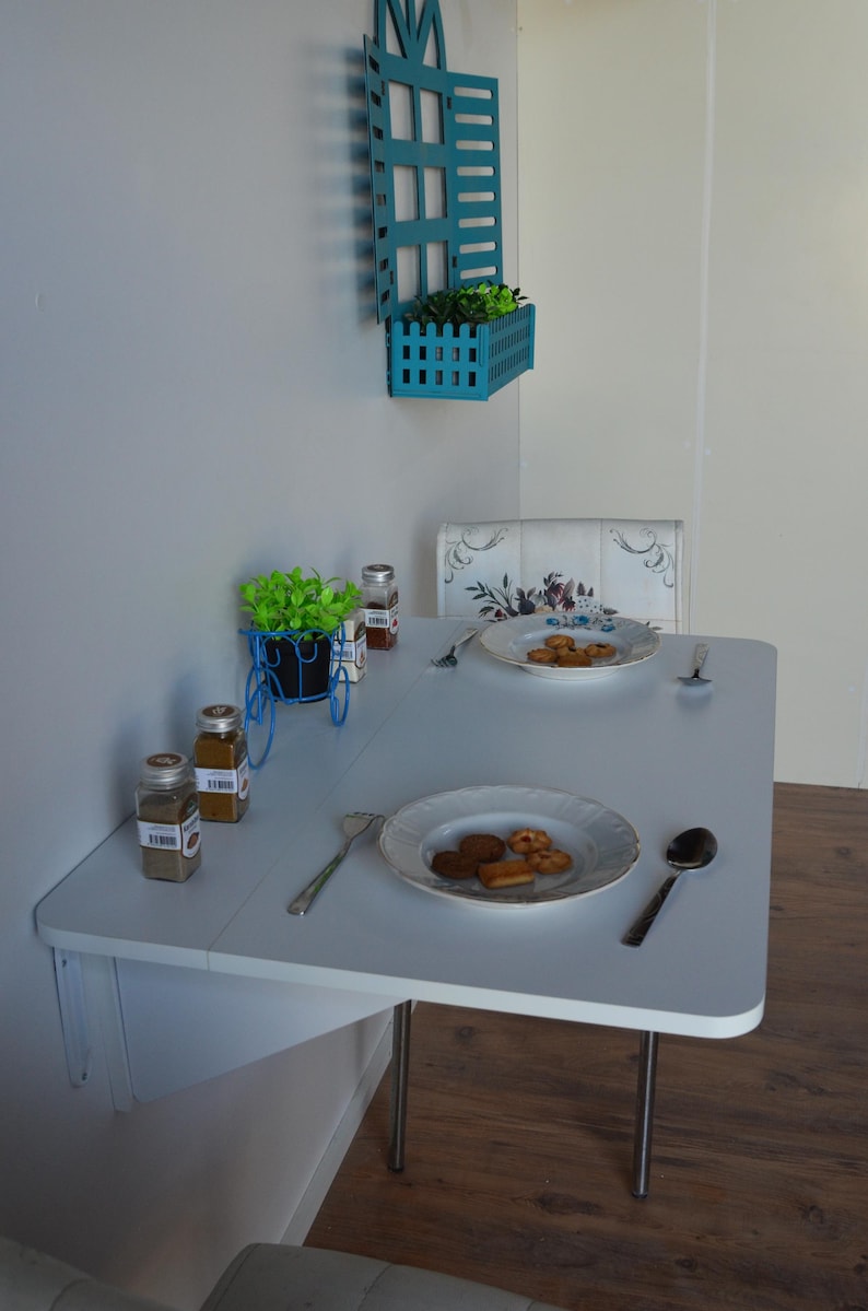 May include: A small, white table with two plates of cookies and silverware. A blue decorative planter with greenery sits on the table, along with spice jars. A blue wall-mounted planter with greenery is above the table.