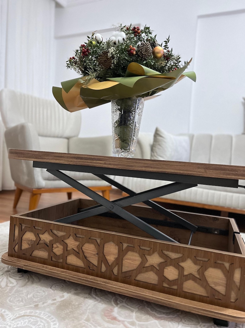 May include: A wooden coffee table with a lift-top mechanism and a storage compartment. The table has a dark brown finish and a geometric pattern on the bottom. The table is raised to a higher level, revealing the storage compartment.