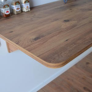 May include: A wooden countertop with a light brown wood grain finish. The countertop is attached to a white surface with metal supports. Several spice jars are on the countertop.
