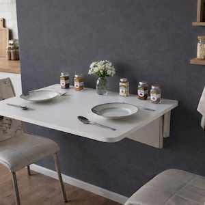 May include: A white, wall-mounted folding table with two plates, spoons, and spice jars. Two chairs with floral patterned backs are positioned on either side. A small vase of white flowers sits in the center.