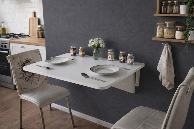 May include: A white, wall-mounted folding table with two plates, spoons, and spice jars. Two chairs with floral patterned backs are positioned on either side. A small vase of white flowers sits in the center.