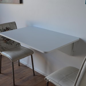 May include: A white, wall-mounted folding table with rounded corners. Two chairs with floral patterned backs and light-colored seats are positioned on either side of the table. The table is approximately 36 inches wide.