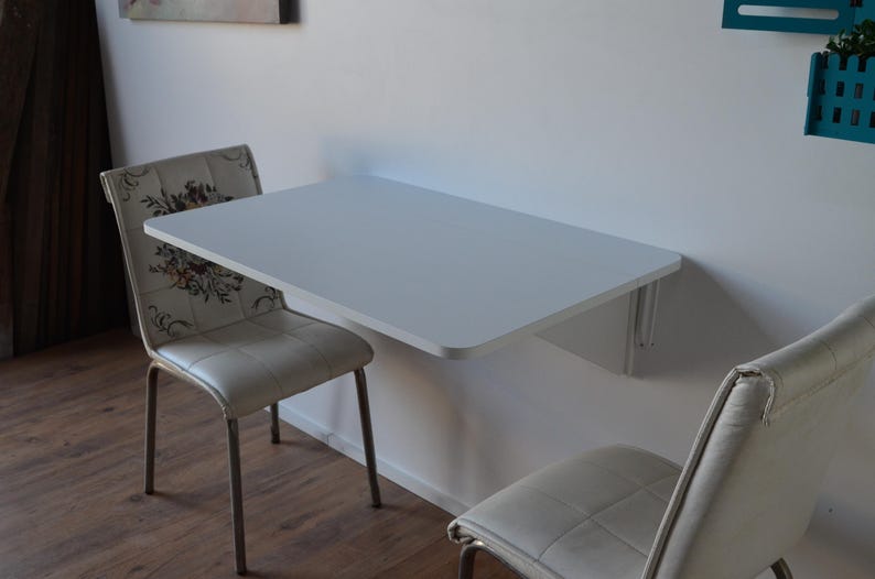 May include: A white, wall-mounted folding table with rounded corners. Two chairs with floral patterned backs and light-colored seats are positioned on either side of the table. The table is approximately 36 inches wide.