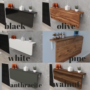 May include: A collection of wall-mounted folding tables in various colors and wood finishes. The tables are shown in black, olive, white, pine, anthracite, and walnut. Each table has a rectangular shape and is mounted on a gray wall.