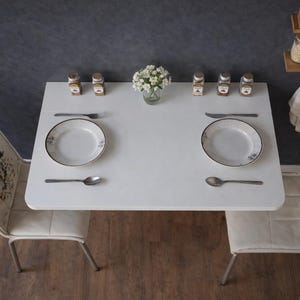 May include: An overhead shot of a small white table set for two. Two chairs with floral designs flank the table. Each place setting includes a plate, spoon, and knife. Small spice jars and a vase of white flowers are on the table.