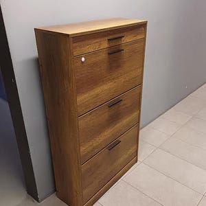 Op de afbeelding: Een bruine houten schoenenkast met vier lades en zwarte metalen handgrepen. De kast heeft een houtnerf afwerking.