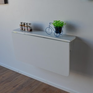 May include: A white, wall-mounted folding table is shown, with a small shelf holding spice jars and a decorative blue bicycle planter with greenery. The table is closed, and the wall is painted white.