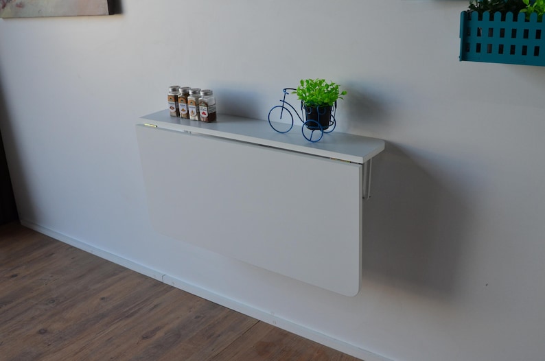 May include: A white, wall-mounted folding table is shown, with a small shelf holding spice jars and a decorative blue bicycle planter with greenery. The table is closed, and the wall is painted white.