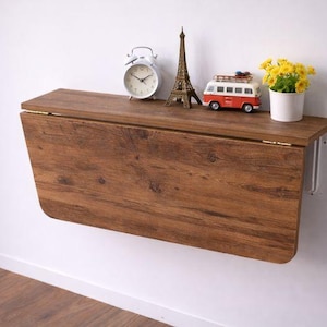 May include: A wall-mounted, folding wooden table with a warm brown finish. The table is adorned with decorative items, including a white alarm clock, a miniature Eiffel Tower, a red and white toy van, and a small pot of yellow flowers.