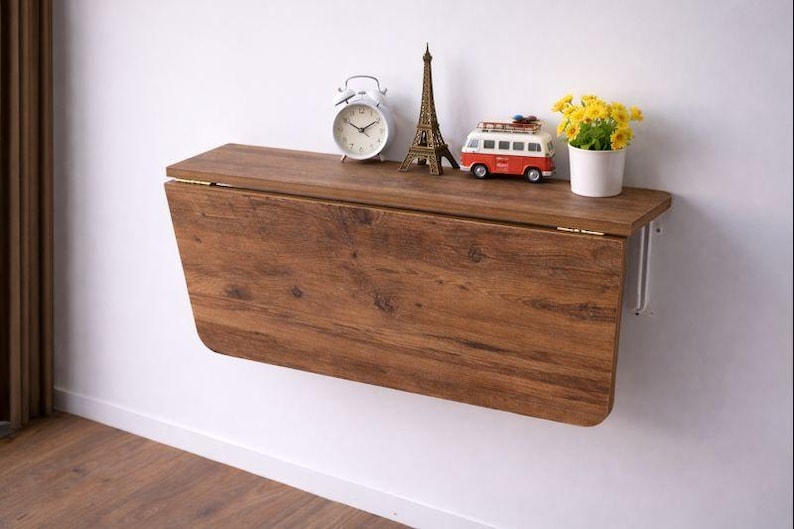 May include: A wall-mounted, folding wooden table with a warm brown finish. The table is adorned with decorative items, including a white alarm clock, a miniature Eiffel Tower, a red and white toy van, and a small pot of yellow flowers.