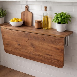 May include: A wall-mounted, folding wooden table with a warm, brown finish. The table is surrounded by a white tiled wall and a white countertop. On the table are a bowl of lemons, a wooden cutting board, a jar, a bottle of oil, and potted plants.