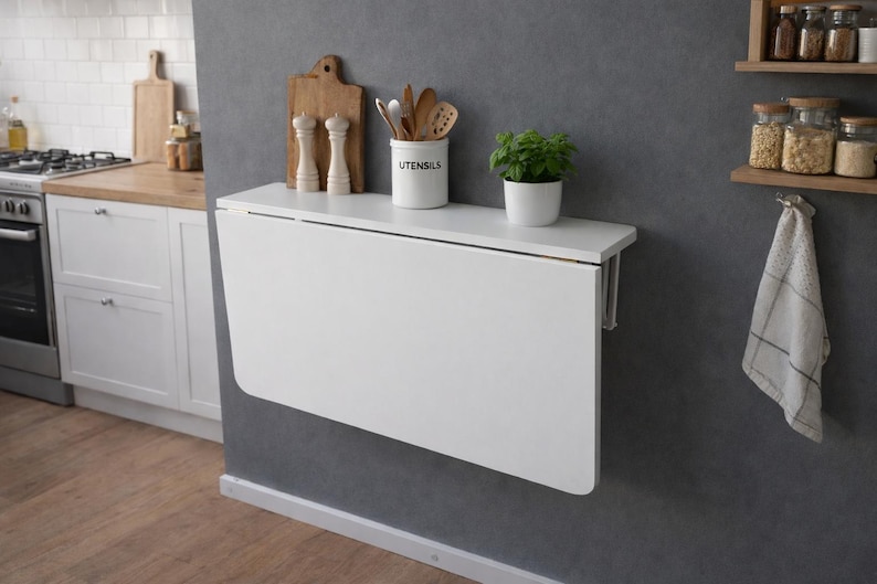 May include: A white, wall-mounted, folding table in a kitchen setting. The table is rectangular and has a clean, modern design. On the table are a small potted plant, a utensil holder, and decorative items. The kitchen has a neutral color scheme.