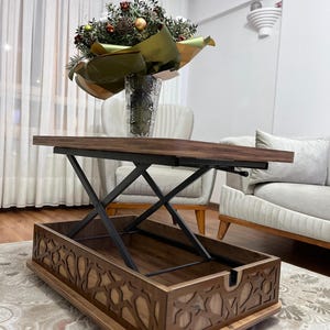 May include: A wooden coffee table with a lift-top mechanism and storage space. The table has a dark brown finish and intricate cutouts around the edges. The table is raised to a higher level, revealing the storage space below.