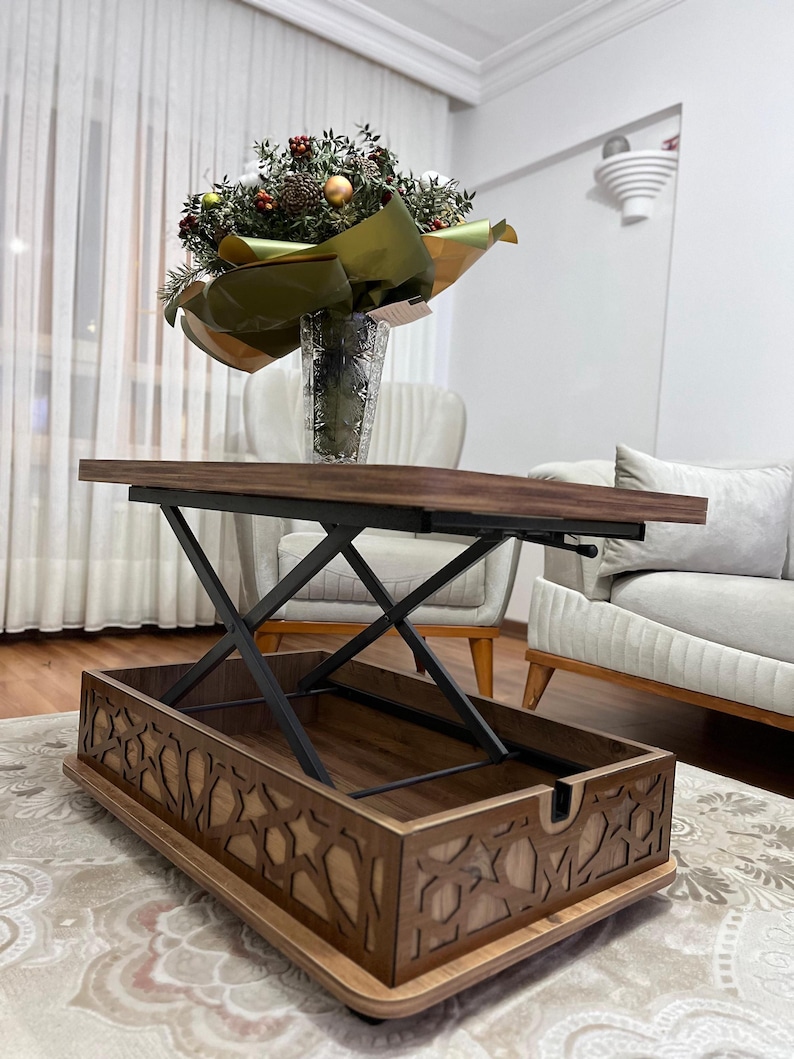 May include: A wooden coffee table with a lift-top mechanism and storage space. The table has a dark brown finish and intricate cutouts around the edges. The table is raised to a higher level, revealing the storage space below.