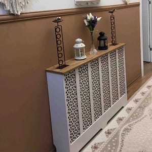 Op de afbeelding: Witte radiatorbekleding met een decoratief geometrisch patroon. De bekleding heeft een houten bovenkant met twee zwarte metalen kaarsenhouders en een kleine witte lantaarn.