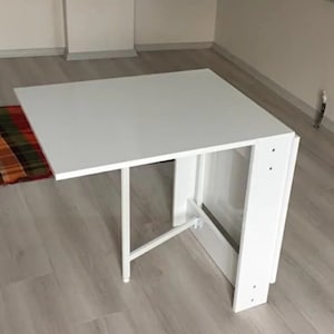 May include: A white folding table with a rectangular top and a white metal frame. The table is folded up and standing on its side.