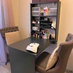 May include: A black table with a white nail lamp on top. There are two brown chairs with pillows. A black shelf unit with multiple shelves is mounted on the wall behind the table. The shelves are filled with bottles of nail polish and other nail care supplies.