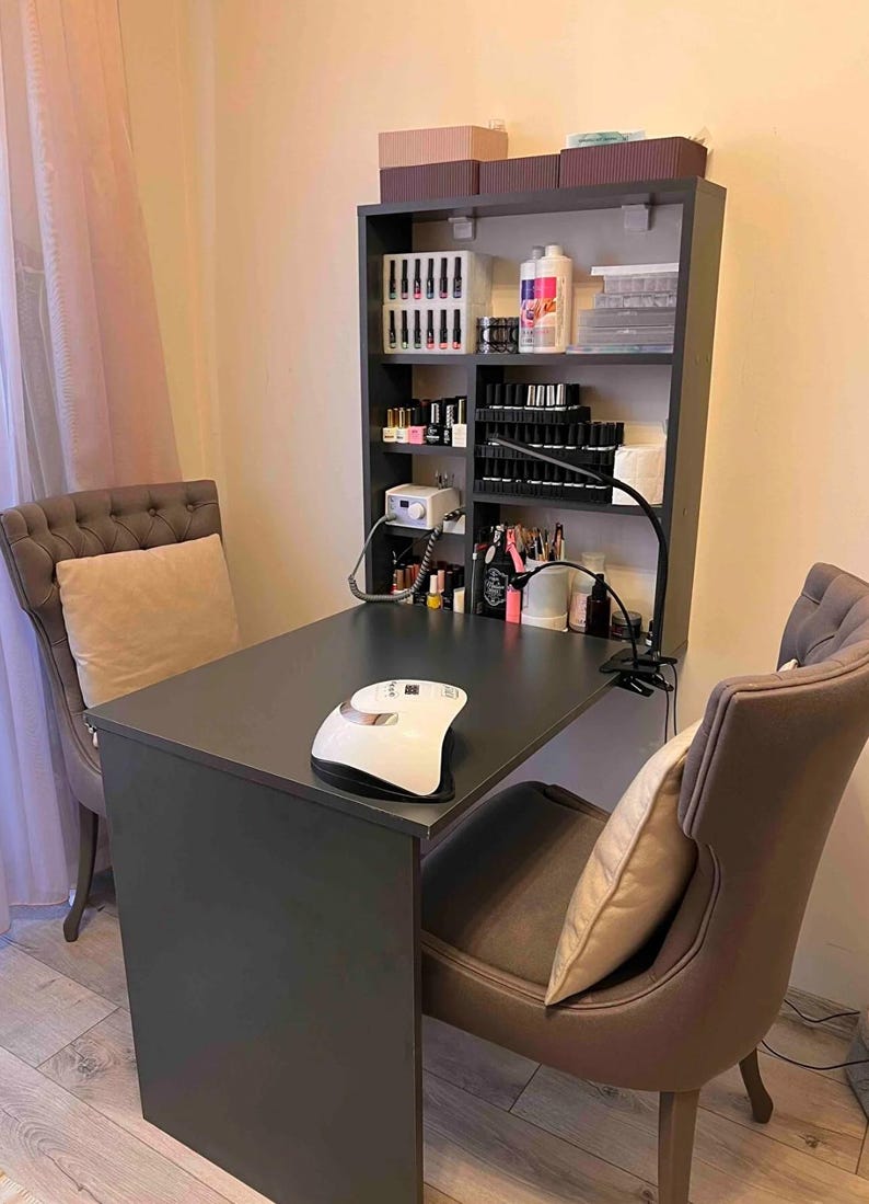 May include: A black table with a white nail lamp on top. There are two brown chairs with pillows. A black shelf unit with multiple shelves is mounted on the wall behind the table. The shelves are filled with bottles of nail polish and other nail care supplies.
