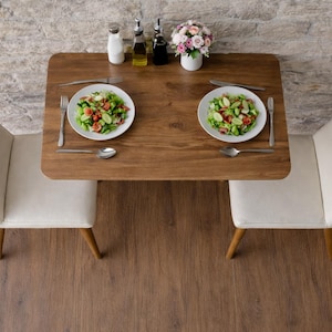 May include: A top-down view of a small dining table set for two. The wooden table has two white plates of salad, silverware, and a small vase of flowers. Two white chairs with wooden legs are positioned on either side of the table.