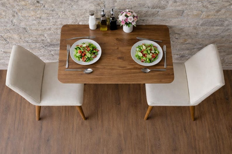 May include: A top-down view of a small dining table set for two. The wooden table has two white plates of salad, silverware, and a small vase of flowers. Two white chairs with wooden legs are positioned on either side of the table.