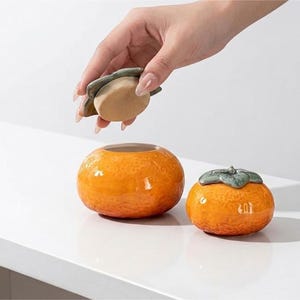 Puede incluir: Dos recipientes de cerámica con forma de caqui. Uno está abierto y el otro tiene tapa. Los recipientes son de color naranja con acabado brillante. La tapa es verde y tiene forma de hoja de caqui. Sobre una superficie blanca.