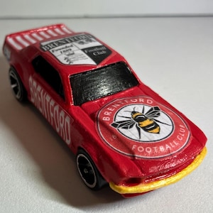 May include: A red toy car with black wheels and a yellow front bumper. The car features the text "Brentford" and the Brentford Football Club logo. The top of the car has white stripes.