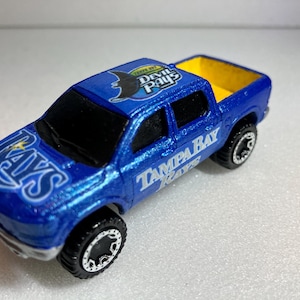 May include: A blue toy pickup truck with the Tampa Bay Rays logo on the hood and sides. The truck bed is yellow. The wheels are black with silver rims. The text "Tampa Bay Rays" is visible on the side.