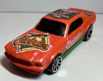 May include: Orange toy car with black windows and silver wheels. The car features the text "Orioles" and a logo on the hood, with "World Series Champions" and the years 1966, 1970, and 1983 on the roof.