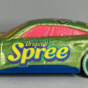 May include: A green toy car with a pink window and a yellow and blue "Original Spree" logo. The car has green wheels and a purple trim.