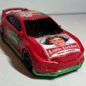 May include: A red toy car with black windows and wheels. The car features the Little Debbie logo on the hood and side, with green accents on the front and side. The car is a miniature replica.
