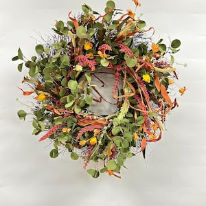 Puede incluir: Una corona floral verde, naranja y roja con hojas de eucalipto y ramas de bayas.