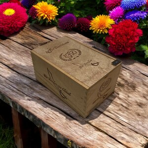 May include: A wooden box with engraved text and designs, including the words "La Flor Dominicana" and "Sir Bender-20 Chisel." The box is light brown and sits on a weathered wooden surface, surrounded by colorful flowers.
