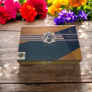 Puede incluir: Una caja de madera con un dise&ntilde;o dorado y azul. La caja tiene el texto "AVO UVEZIAN" y "SYNCHRO SOUTH AMERICA" en ella. La caja est&aacute; cerrada y tiene un cierre de metal.
