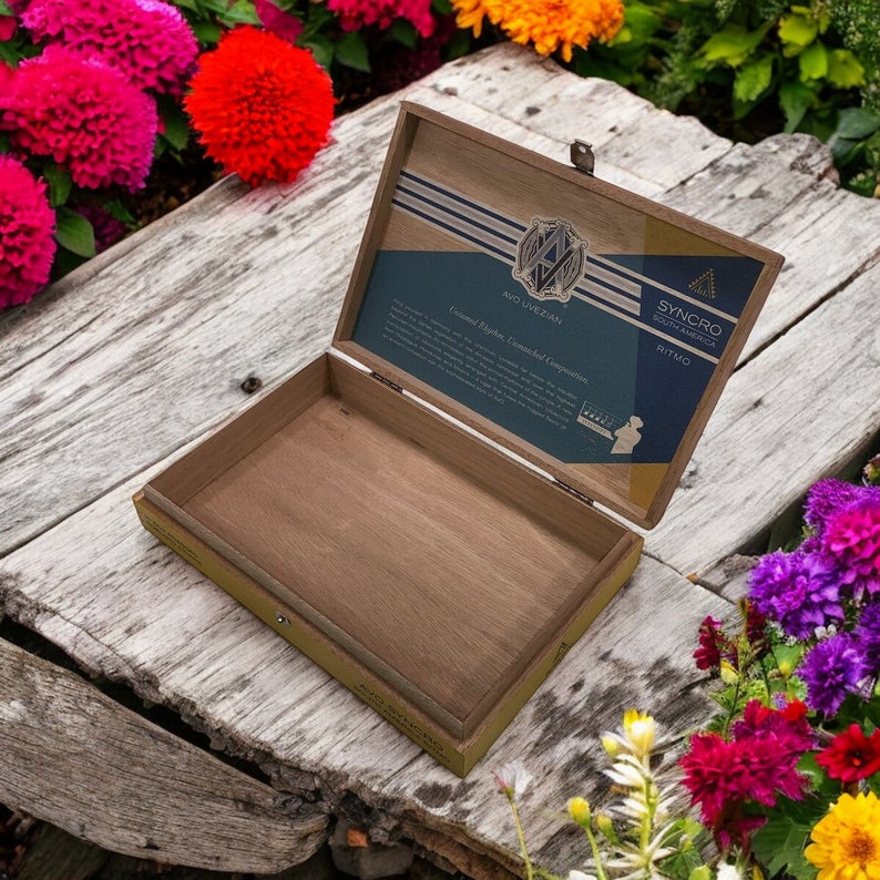 Puede incluir: Una caja de madera con tapa abatible, abierta para revelar un interior de madera natural. La tapa presenta un dise&ntilde;o impreso con el texto "AVO IVEZIAN", "SYNCHRO SOUTH AMERICA" y "RITMO".