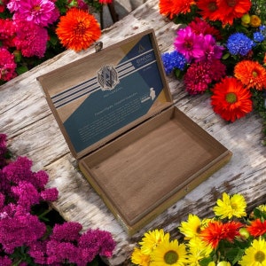 Puede incluir: Una caja de madera con tapa abatible, abierta para revelar un interior marr&oacute;n. La tapa presenta una etiqueta azul y dorada con el texto "SYNCHRO SOUTH AMERICA" y "A&Ntilde;O 2022". La caja est&aacute; rodeada de flores de colores.