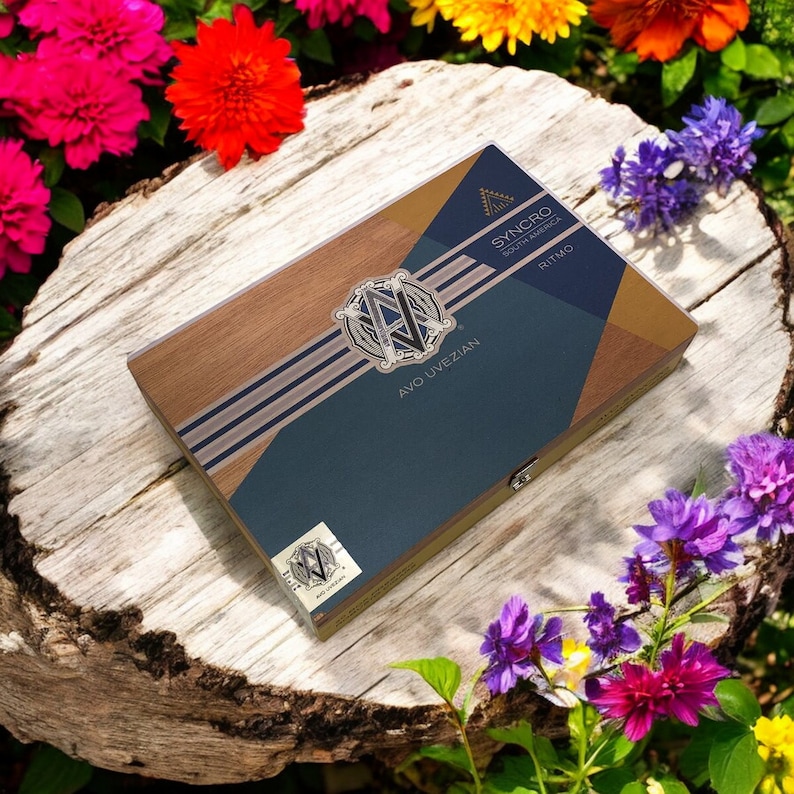 Puede incluir: Una caja de madera con un dise&ntilde;o azul y dorado. La caja tiene el texto "AVO UVEZIAN" y "SYNCHRO SOUTH AMERICA" en ella. La caja est&aacute; sentada sobre una superficie de madera con flores alrededor.