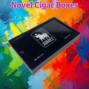 May include: Black cigar box with the word "JUAREZ" and a bull logo in white. The box also has the text "12 CIGARS - 4 X 50" and "MINI DOZER LE 2026". The words "Novel Cigar Boxes" are at the top.