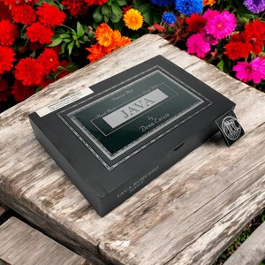 Op de afbeelding: Een zwarte houten doos met een zilveren label waarop "Java by Drew Estate" en "Premium Mix - Long Filler - Hand Made" staat. De doos staat op een houten oppervlak met bloemen op de achtergrond.