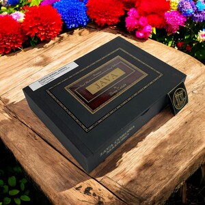 Op de afbeelding: Een zwarte sigarenkist met gouden letters en accenten, met het woord "JAVA" prominent weergegeven. De kist is gelabeld met "JAVA CORONA MADURO" en "Drew Estate". De kist staat op een houten oppervlak, omringd door kleurrijke bloemen.